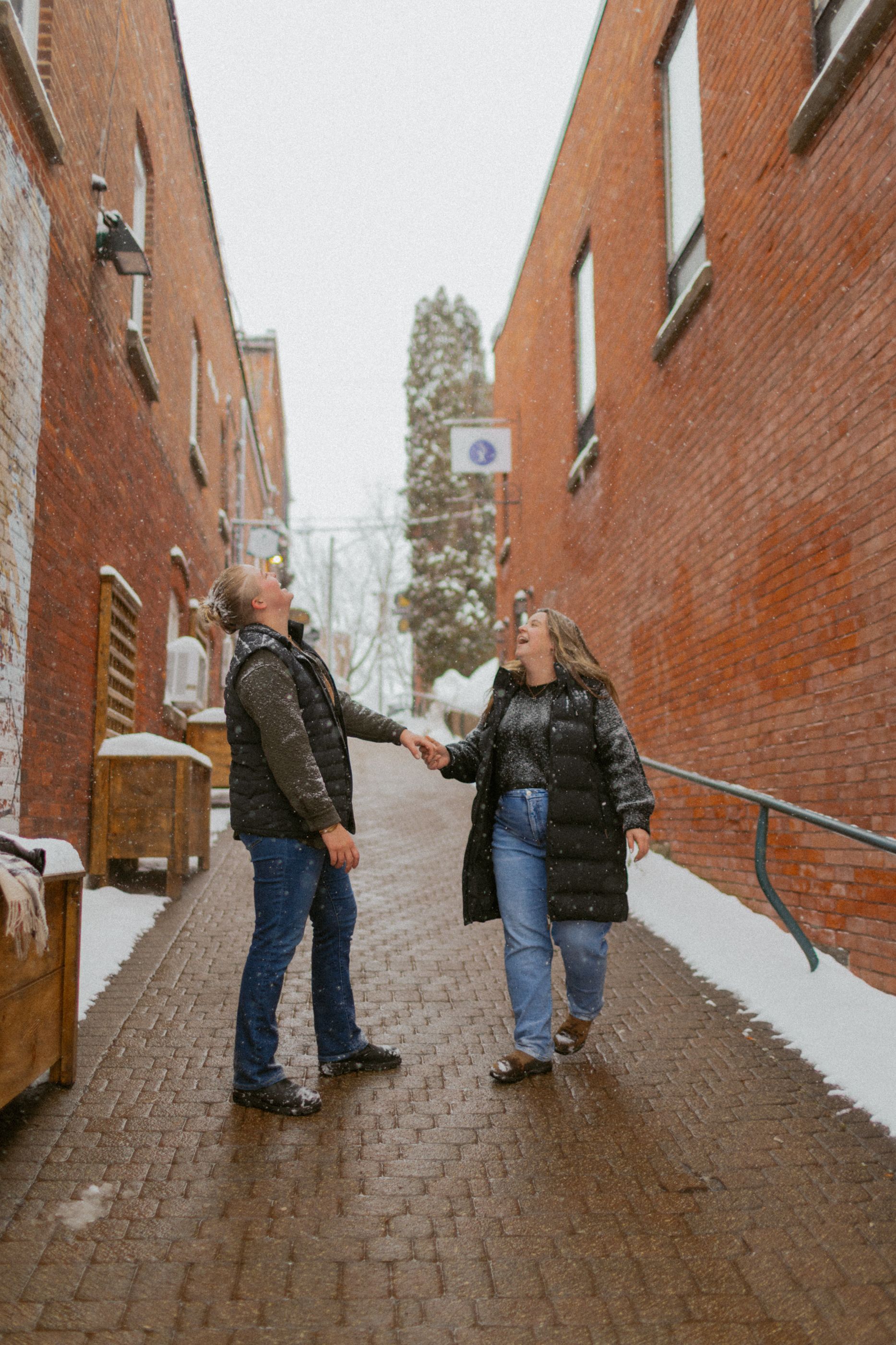 Two people hold hands while walking down a snowy brick alleyway between buildings.