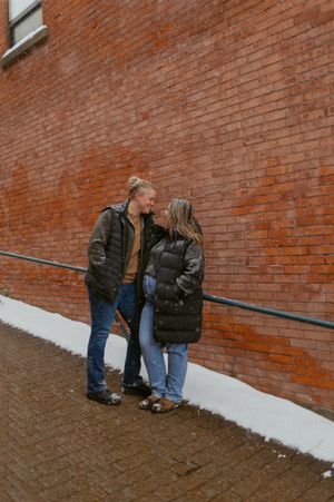 A couple in winter coats share an intimate moment against a brick wall.