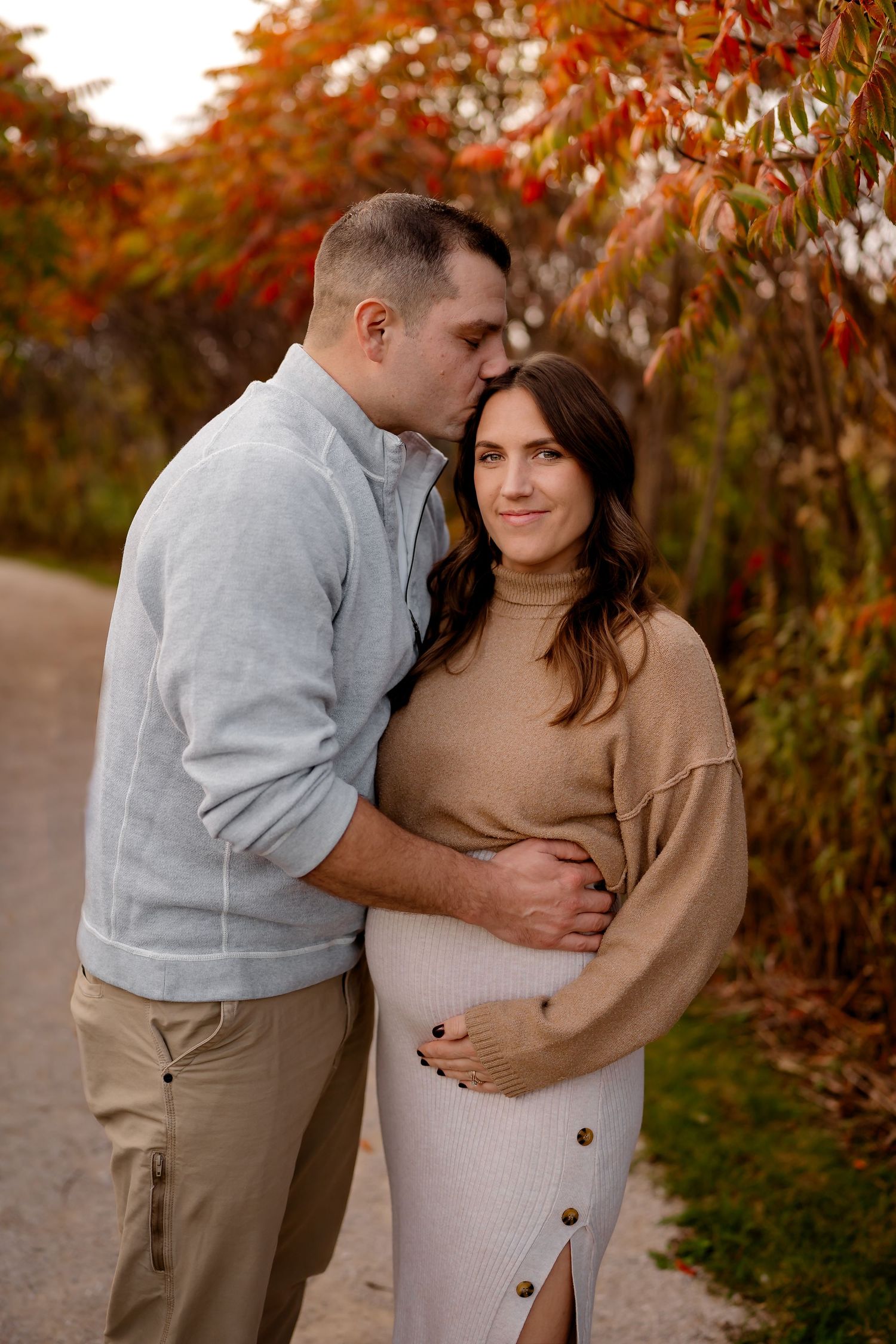 A couple shares an intimate moment among autumn foliage wearing coordinating tan and gray outfits.