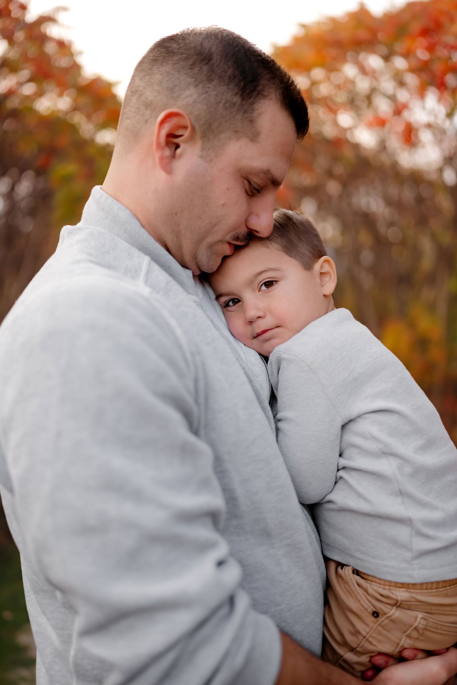 Parent and child wearing matching gray sweatshirts embrace tenderly during autumn photoshoot with orange leaves in background.