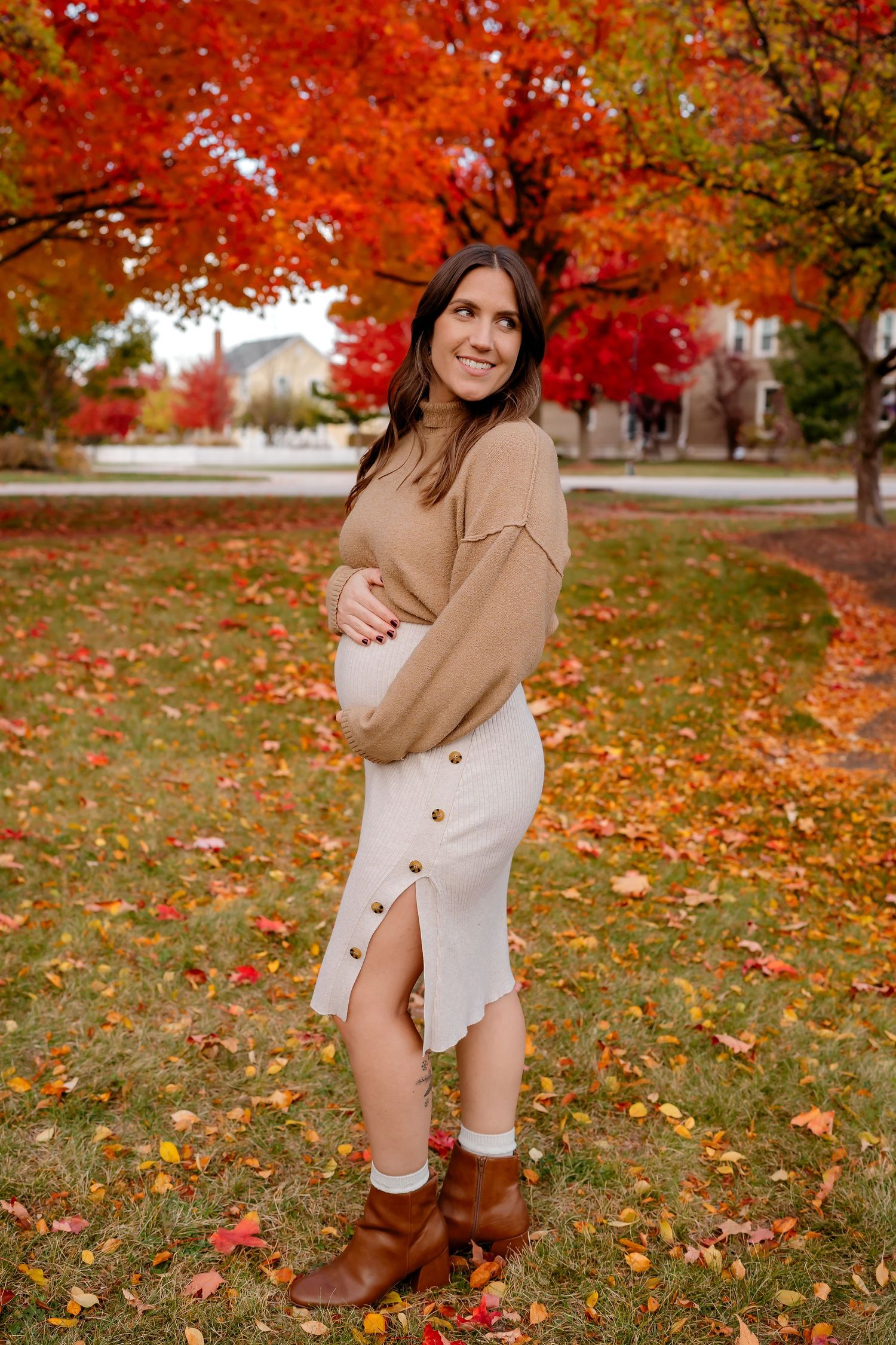 Person in beige sweater poses in autumn setting with vibrant red maple trees and fallen leaves in background.