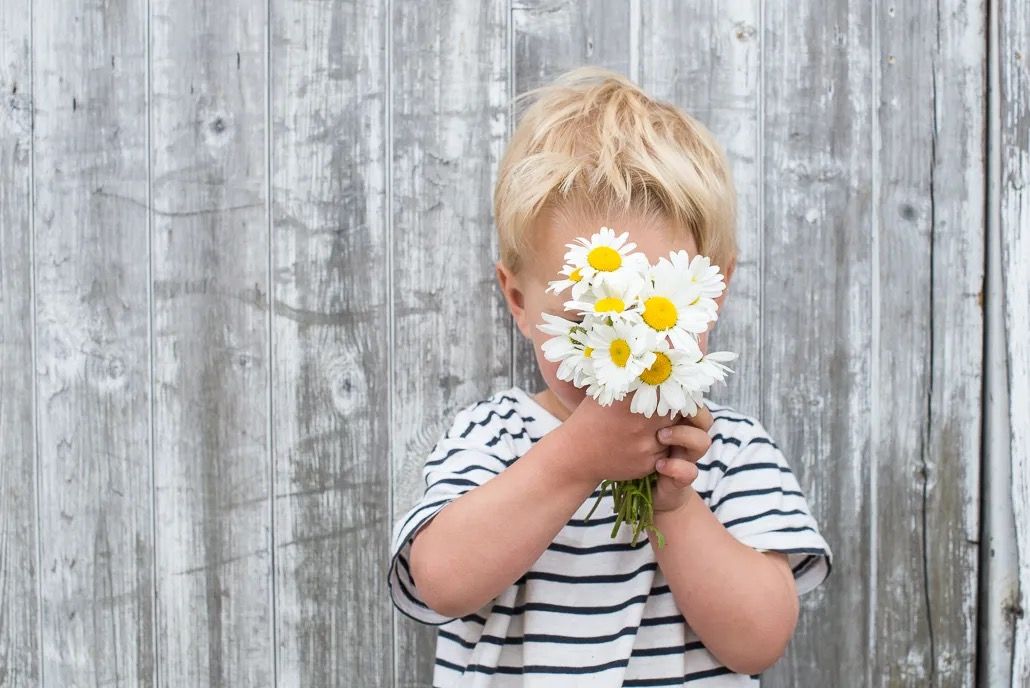 Young child holding a bunch of daisies in front of their face against a wooden backdrop.