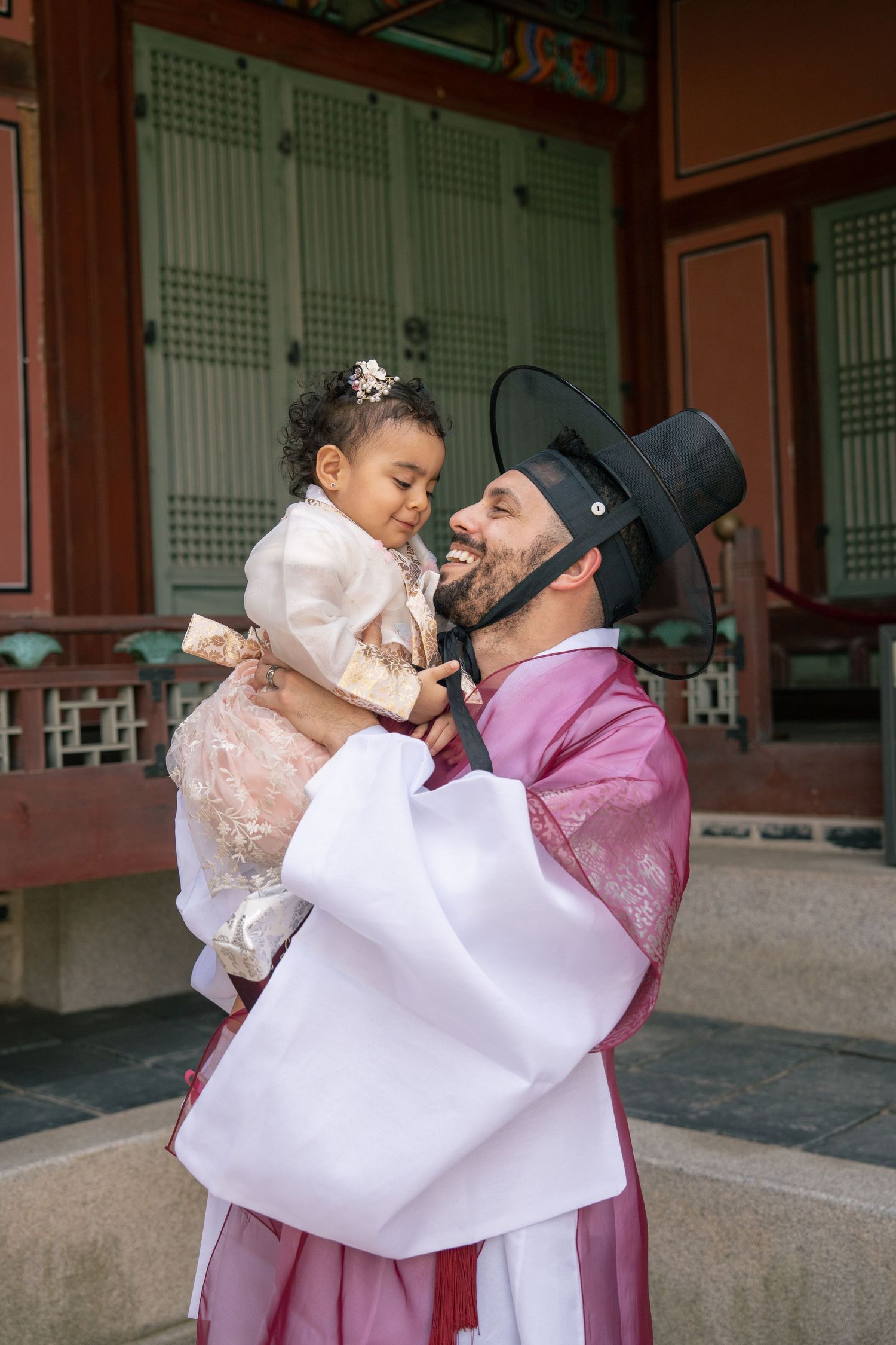 A family wearing traditional Korean hanbok clothing embraces in a tender moment at a historic temple courtyard.