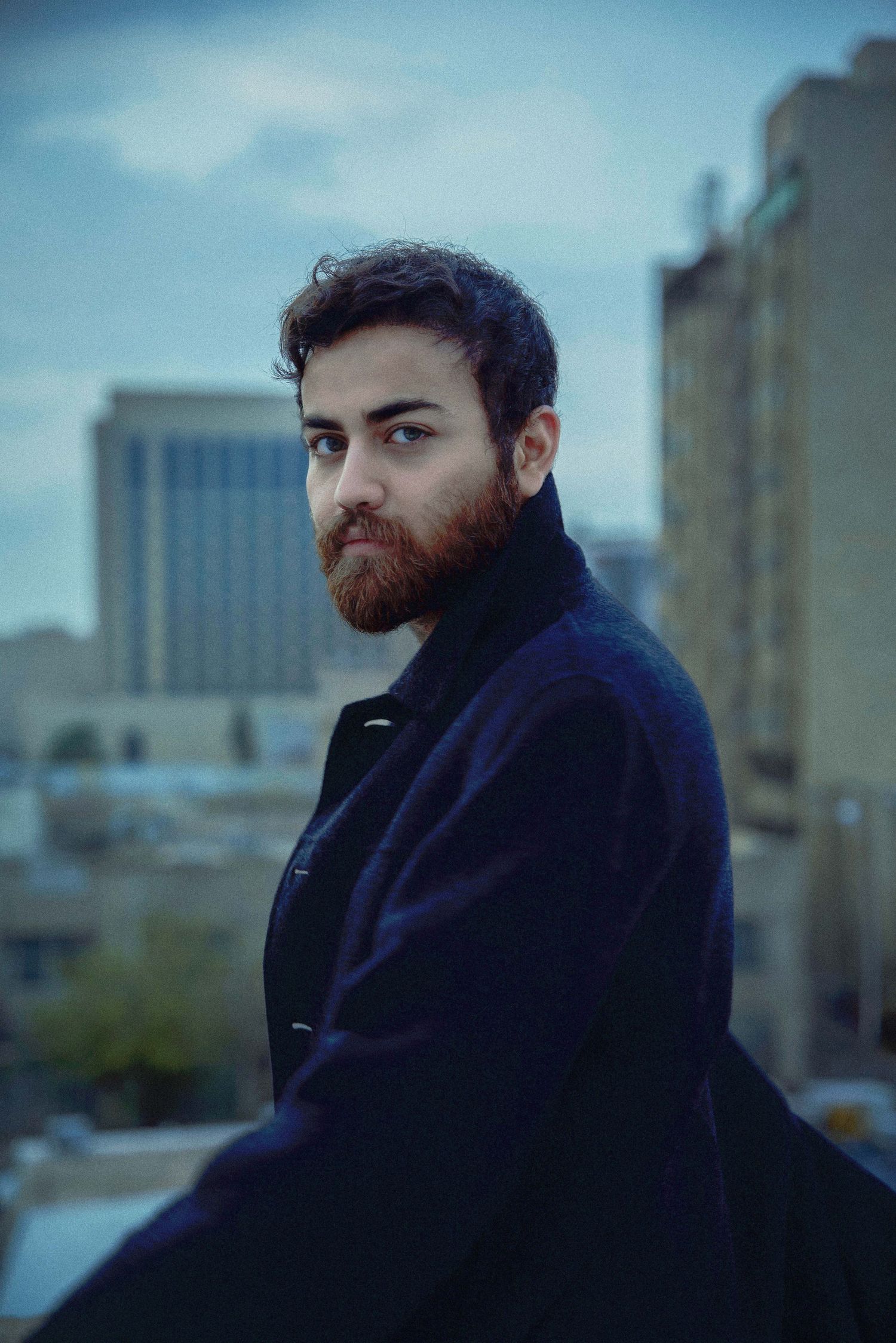 A bearded person in a dark coat looks thoughtfully to the side against an urban cityscape backdrop.