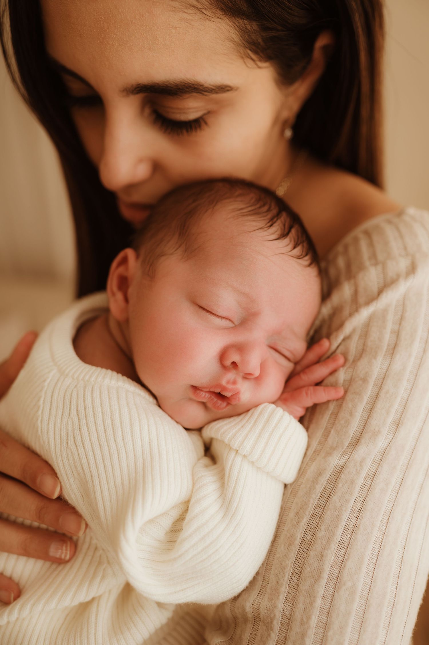 A sleeping newborn baby in a white knit sweater is tenderly cradled against someone wearing cream-colored clothing.