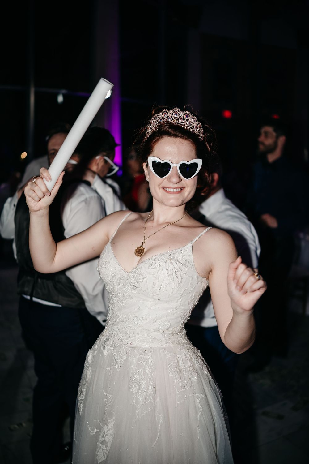 A bride in a white lace dress and sunglasses dances joyfully with glowsticks at a wedding reception.