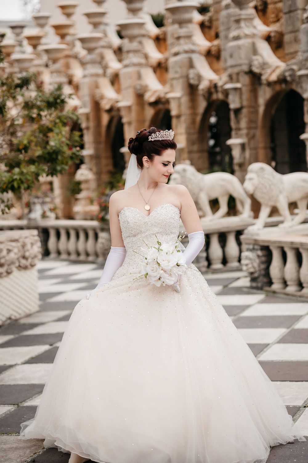 A bride in a voluminous white tulle gown sits gracefully on stone steps of an ornate architectural setting.