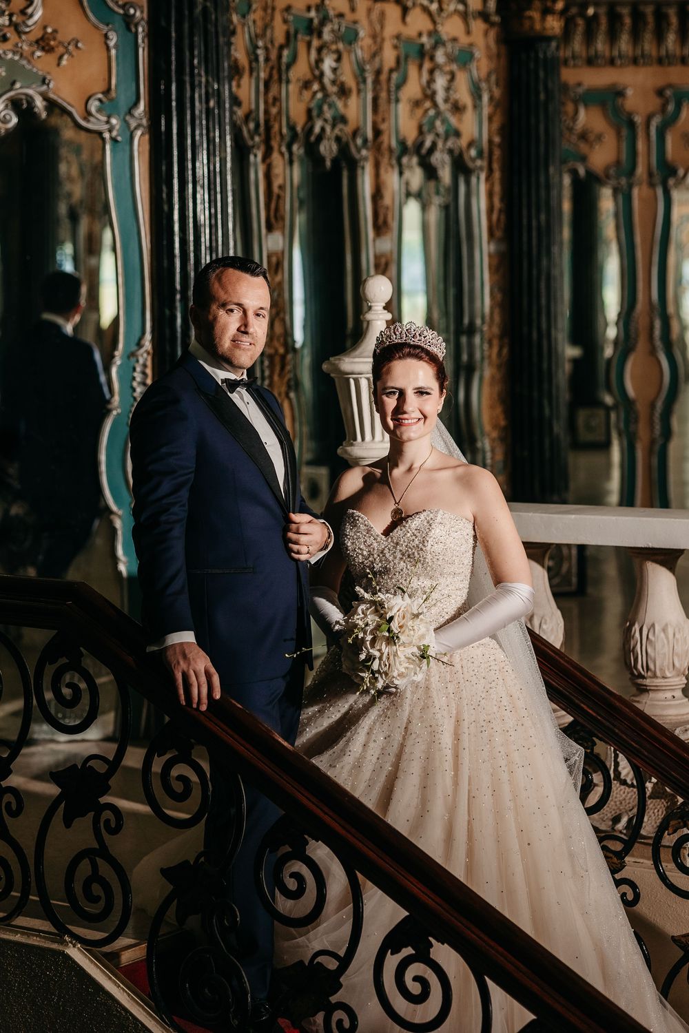 Elegant wedding couple poses on ornate staircase with intricate wrought iron railings in vintage-style venue.