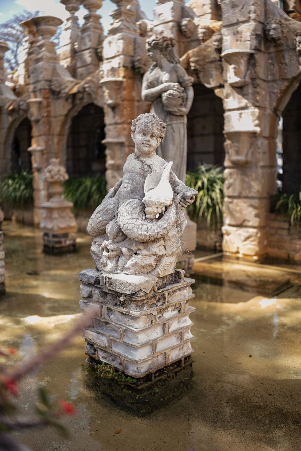 Stone cherub statue holding a book stands in ornate architectural setting with water feature.
