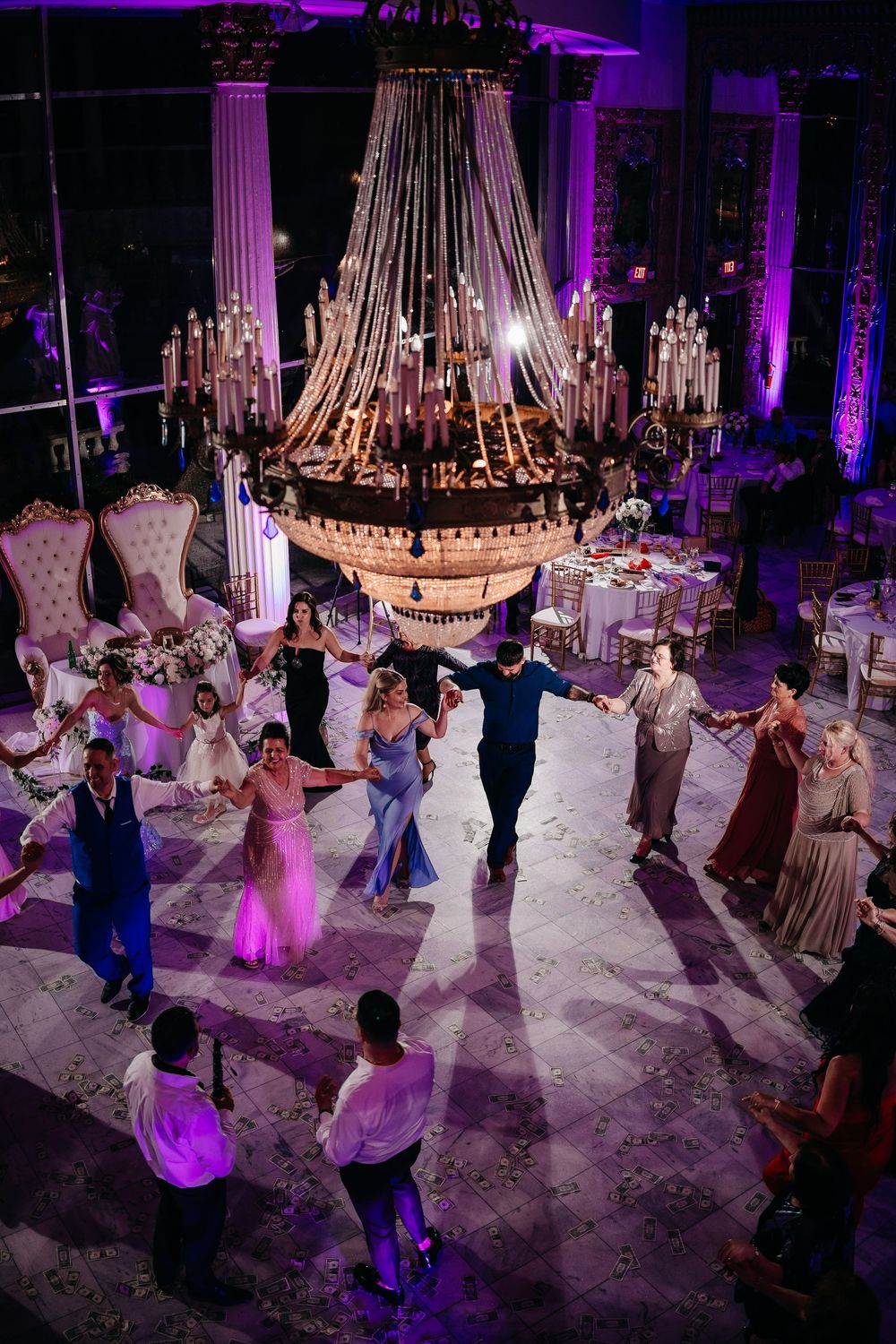 Wedding guests dance on a purple-lit dance floor under elegant chandeliers at an evening reception celebration.
