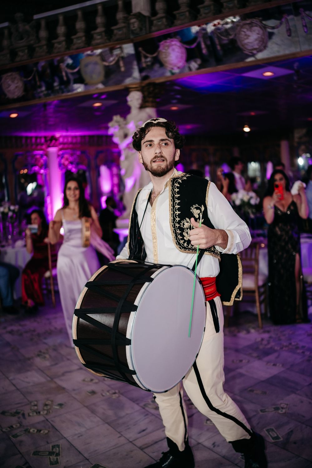Traditional drummer in folk costume performs at an event with purple lighting.
