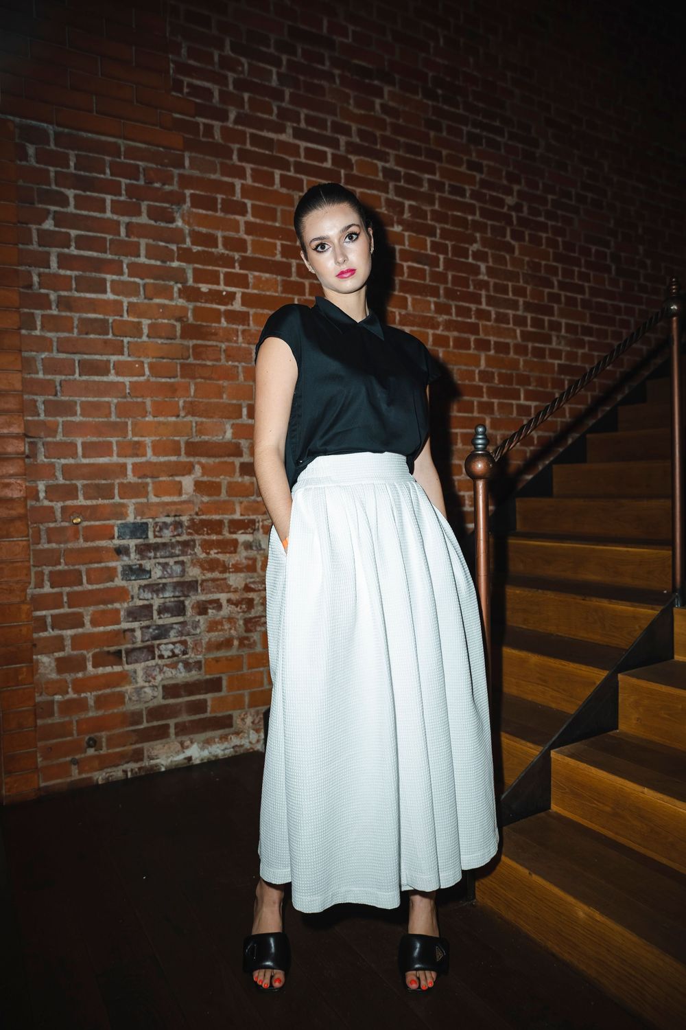 Fashion model wearing black and white dress poses against brick wall on wooden stairs.