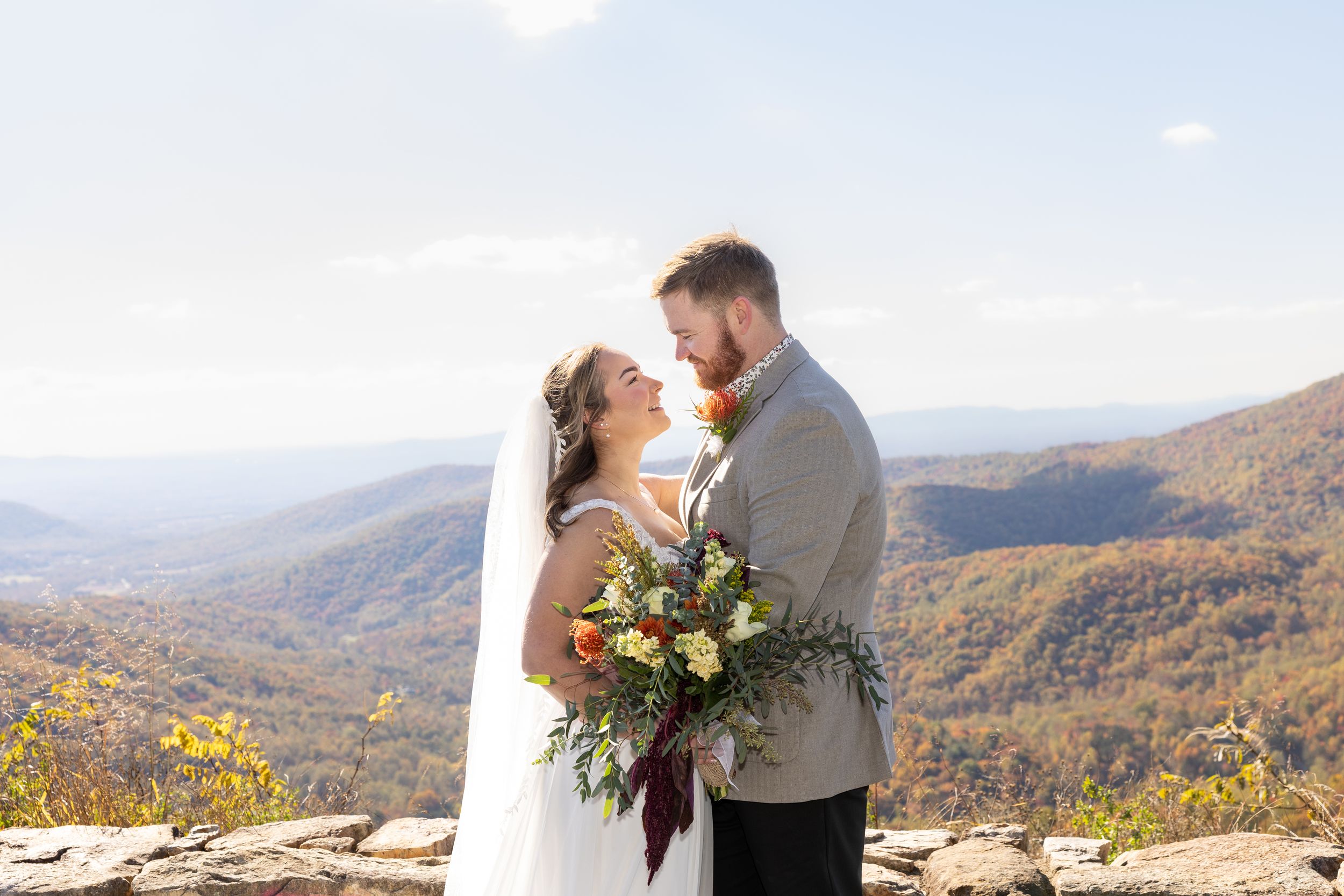 A couple shares a romantic moment on a mountaintop overlook with autumn foliage and rolling hills in the background.