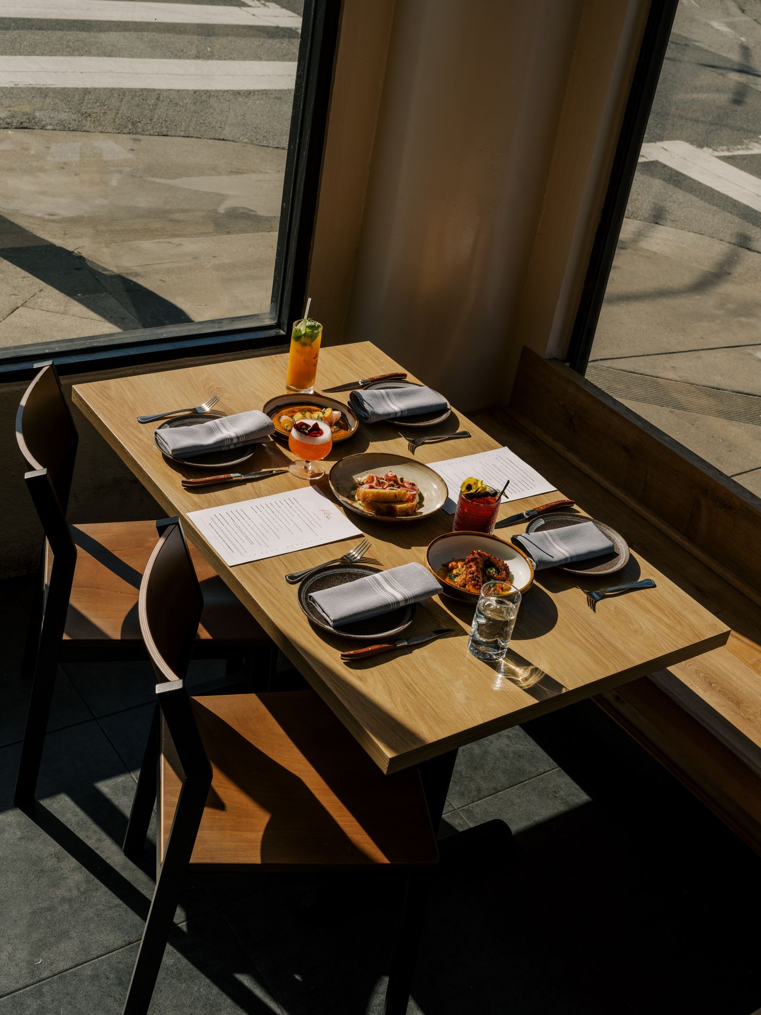 Wooden restaurant table set with place settings and drinks illuminated by natural sunlight through a window.