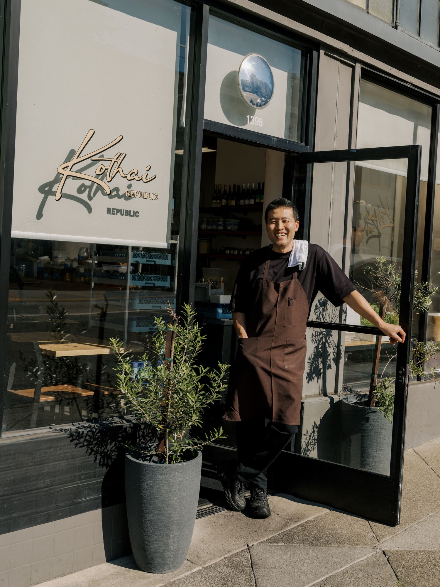 Modern restaurant storefront with large windows, potted plants, and signage visible from the sidewalk.
