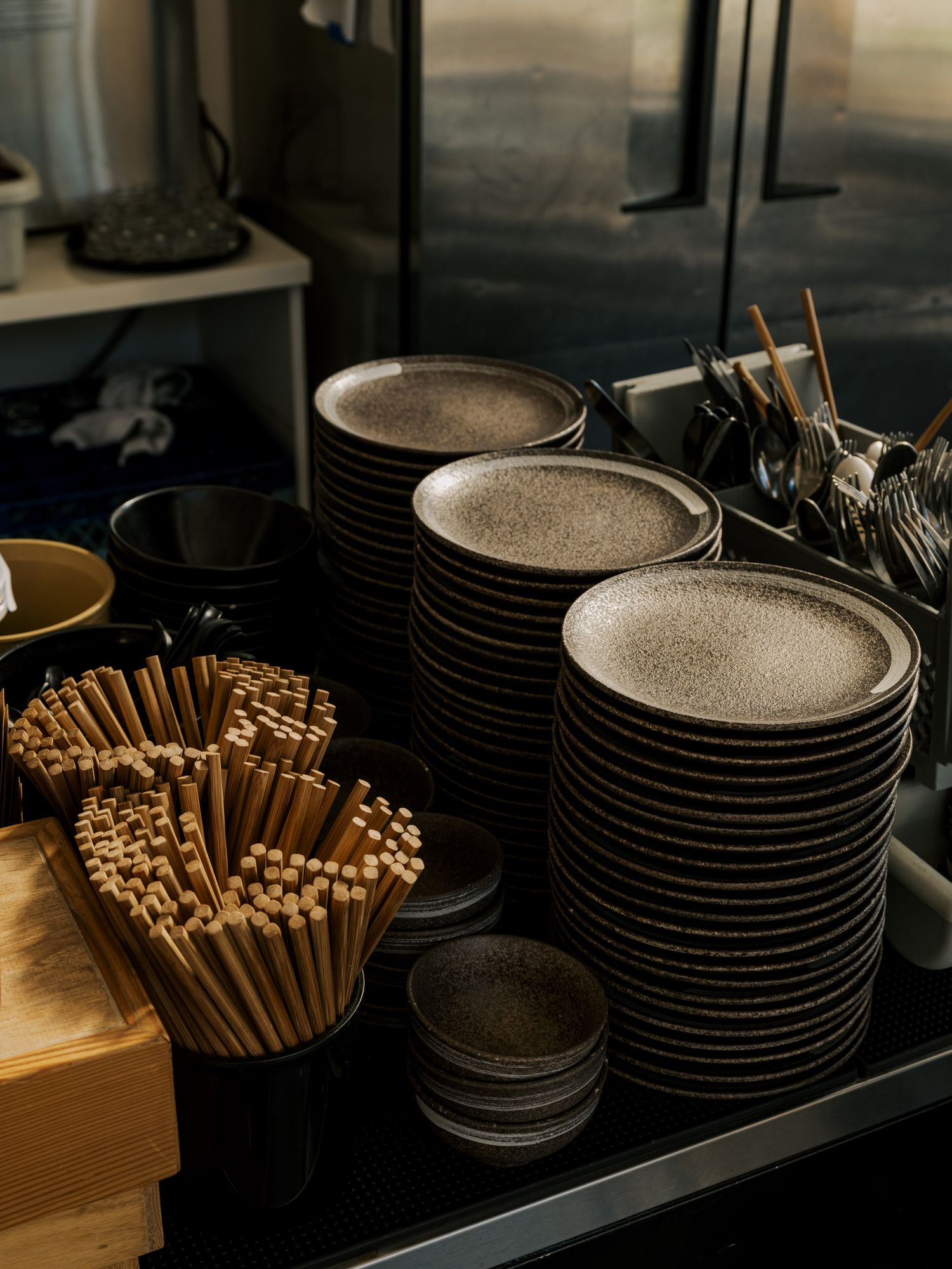 Stacks of gray ceramic plates and wooden chopsticks arranged neatly on dark restaurant shelving.