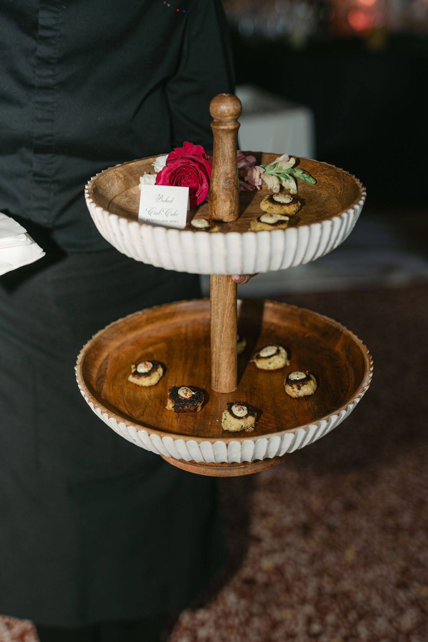 Rustic two-tiered serving stand with fluted edges displays small decorated pastries and cookies on dark background.