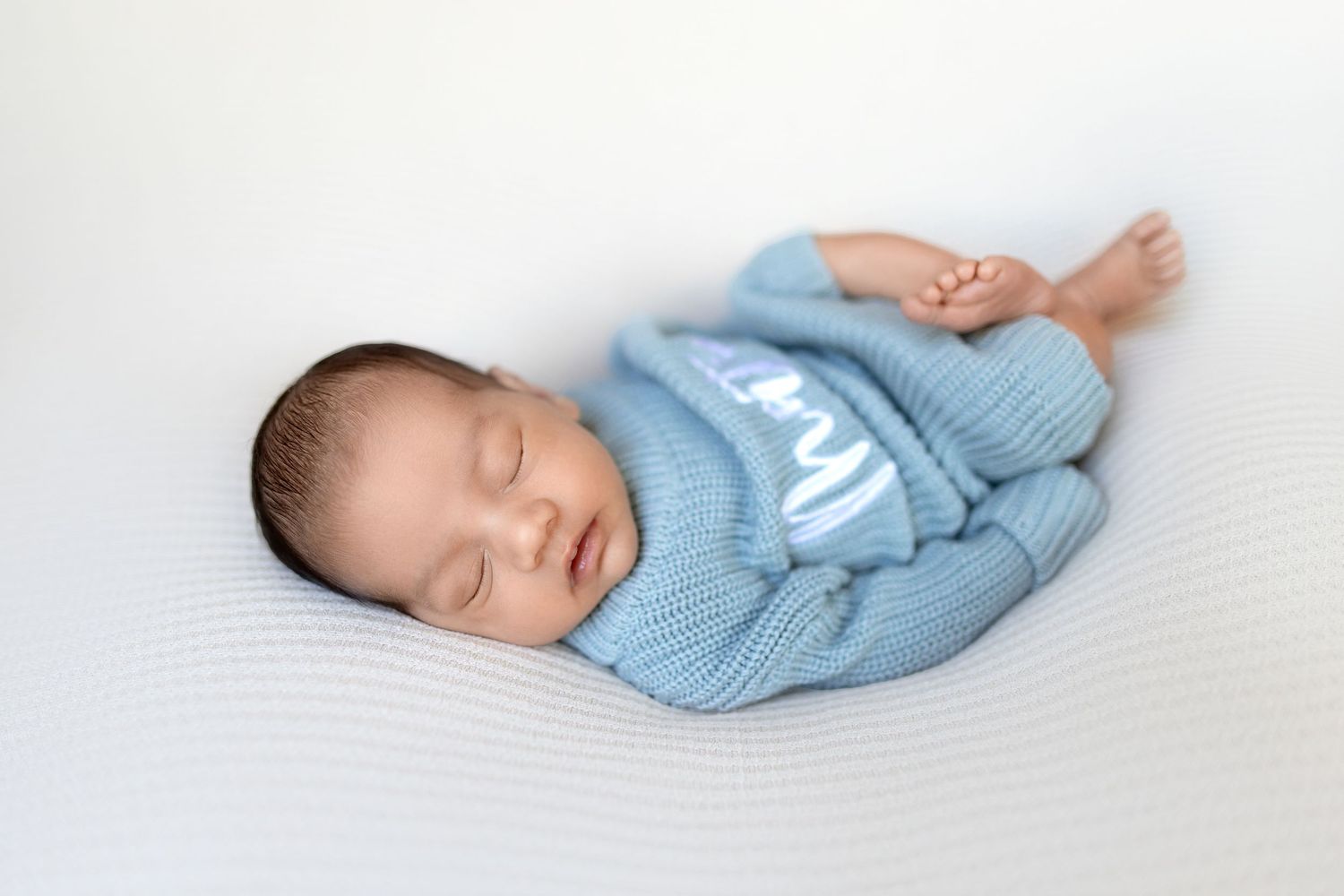 Peaceful sleeping newborn baby wearing soft blue sweater rests on white blanket in natural light.