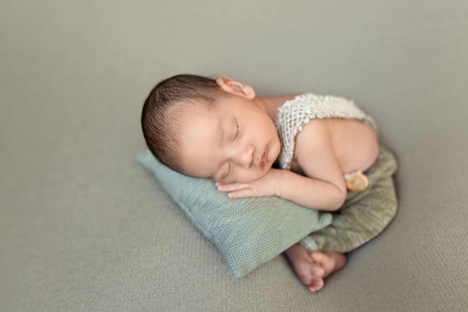 Sleeping newborn baby curled up on soft gray blanket with light blue pillow in professional studio setting.