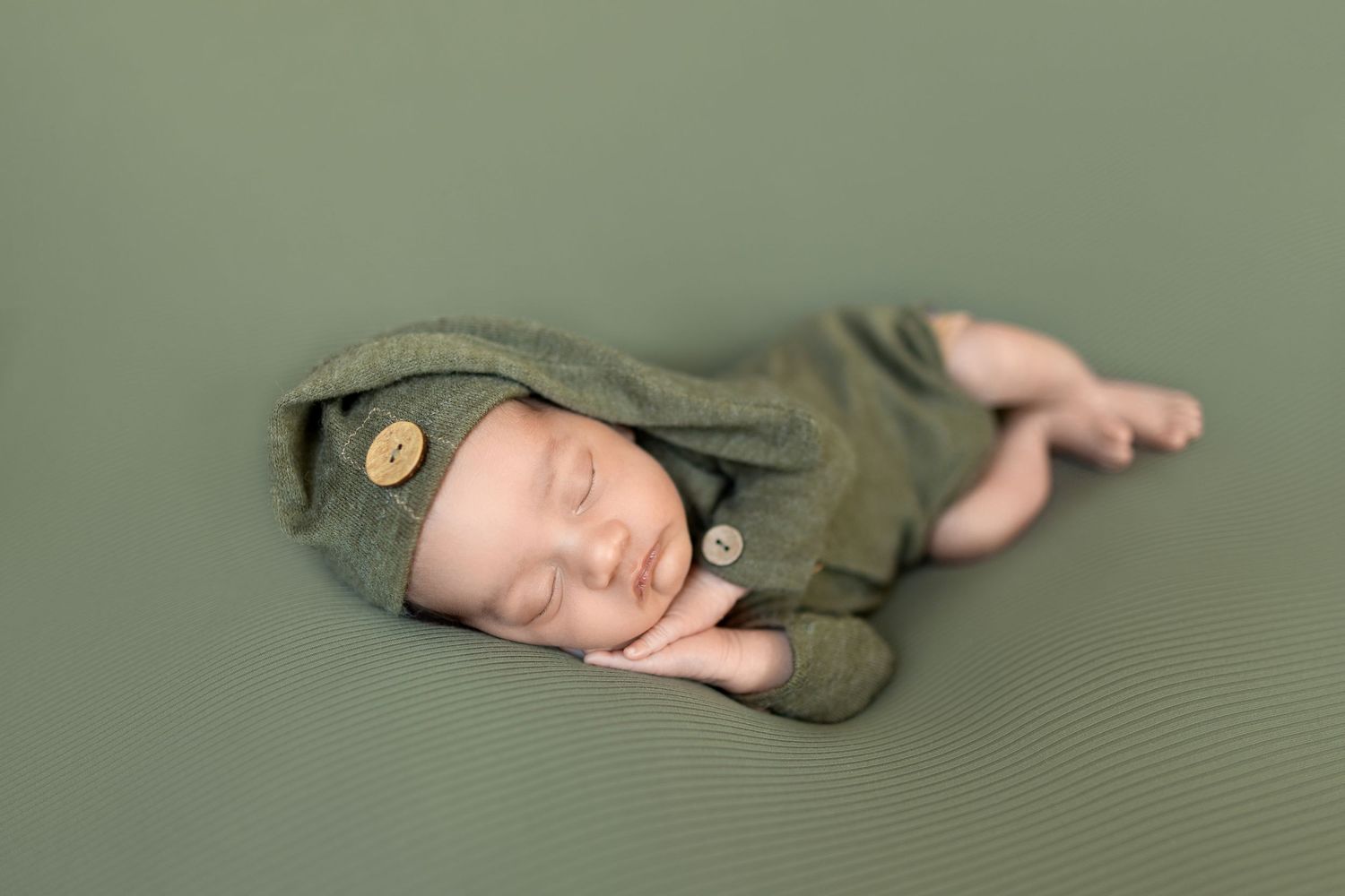 Sleeping newborn baby wrapped in olive green fabric lying on textured gray backdrop during professional photo session.