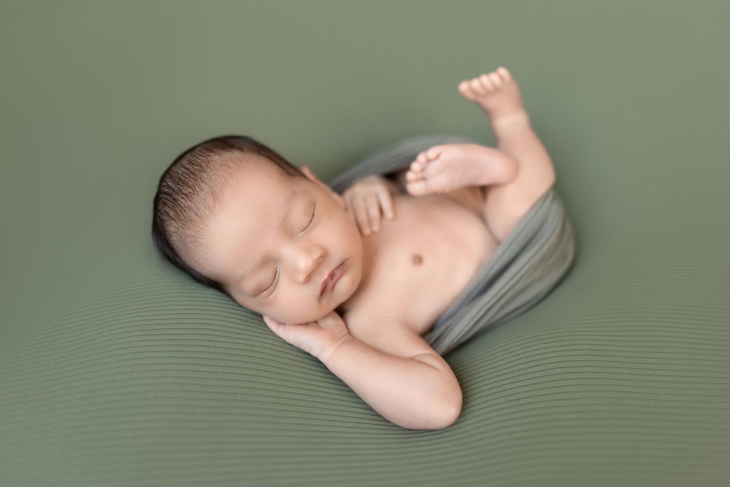 A sleeping newborn baby lies peacefully on a sage green blanket in a curled position.