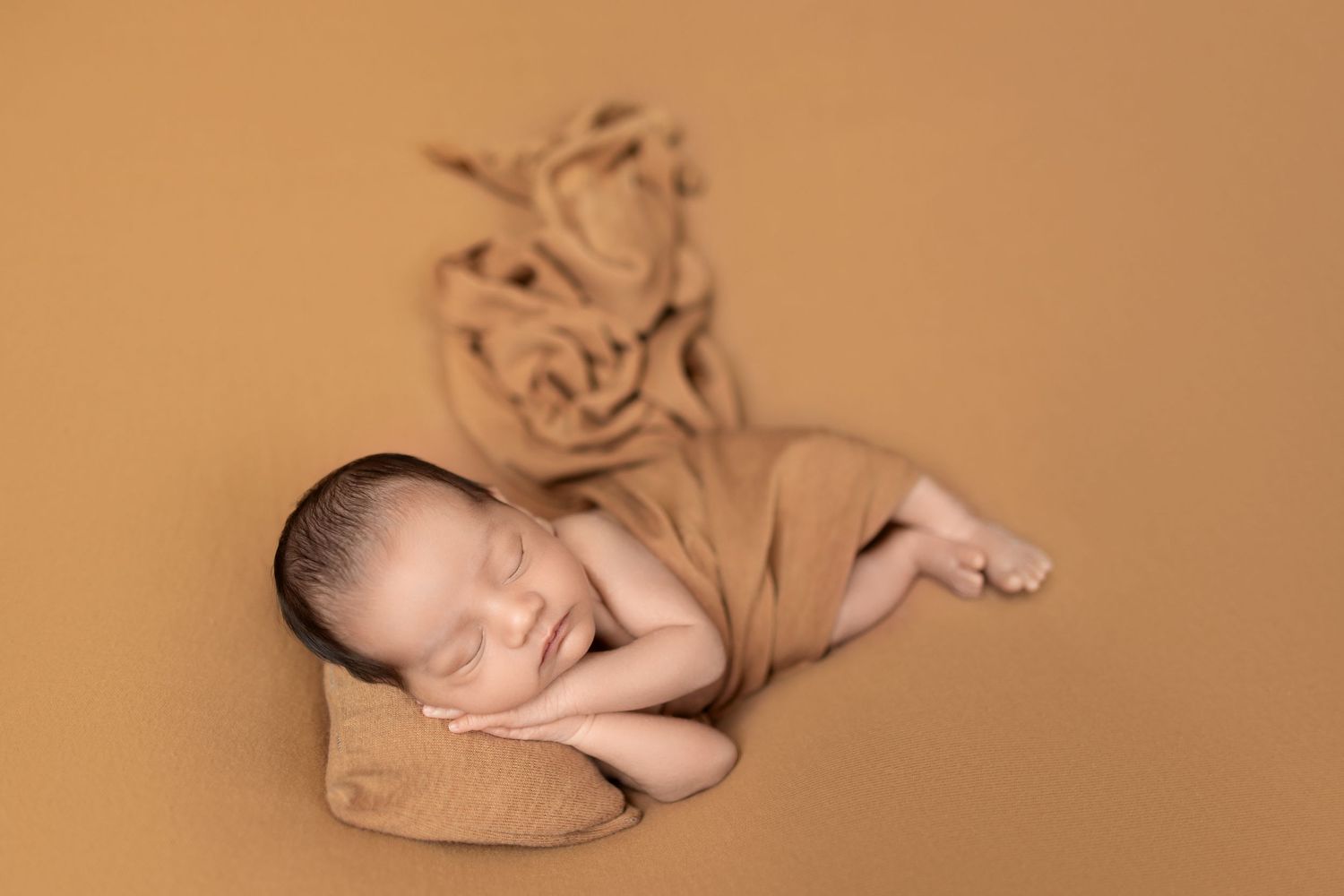 Peaceful newborn baby sleeping on tan fabric against warm brown background during professional photo session.