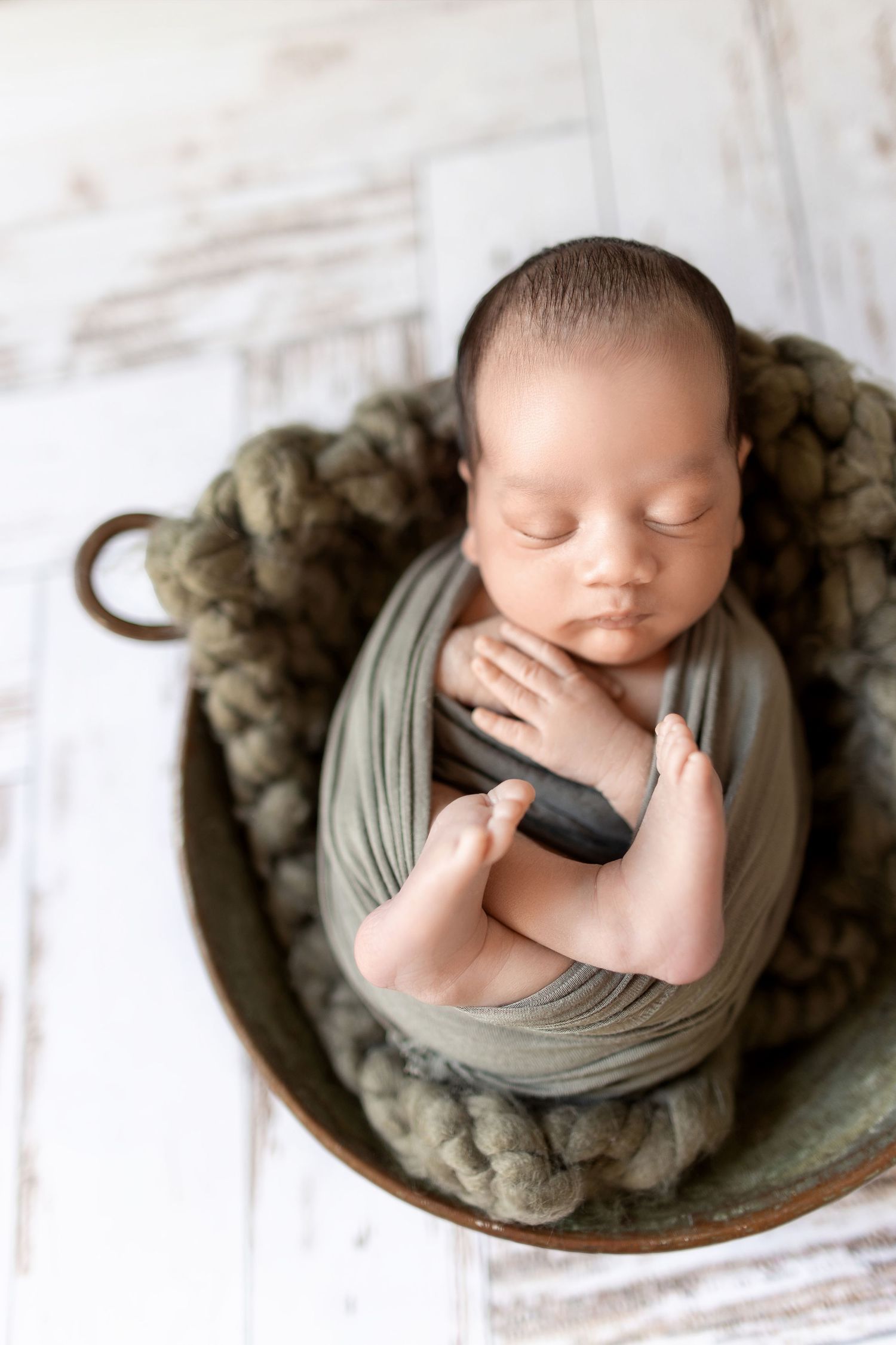 A sleeping newborn wrapped in soft gray fabric lies curled up on a textured round surface in a professional portrait.