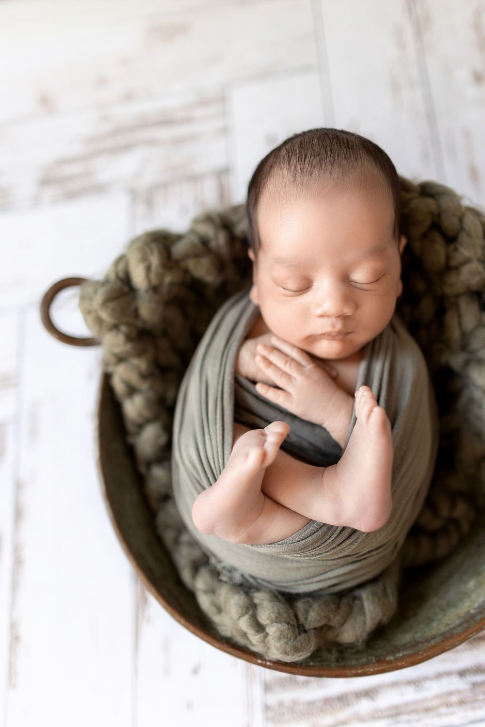 A sleeping newborn wrapped in soft gray fabric lies curled up on a textured round surface in a professional portrait.