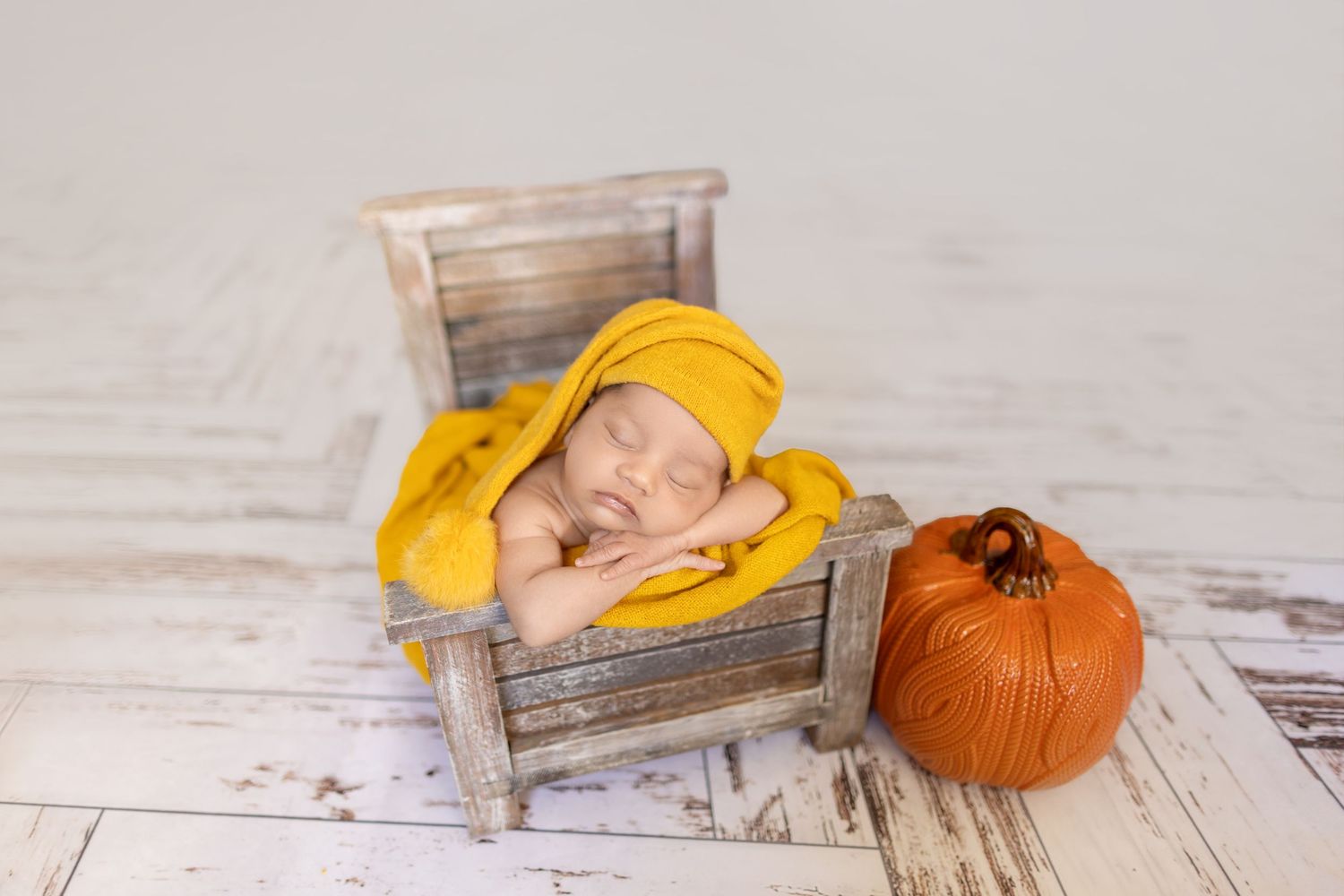 A baby in a yellow knit hat lies sleeping on a rustic wooden surface next to an orange pumpkin.