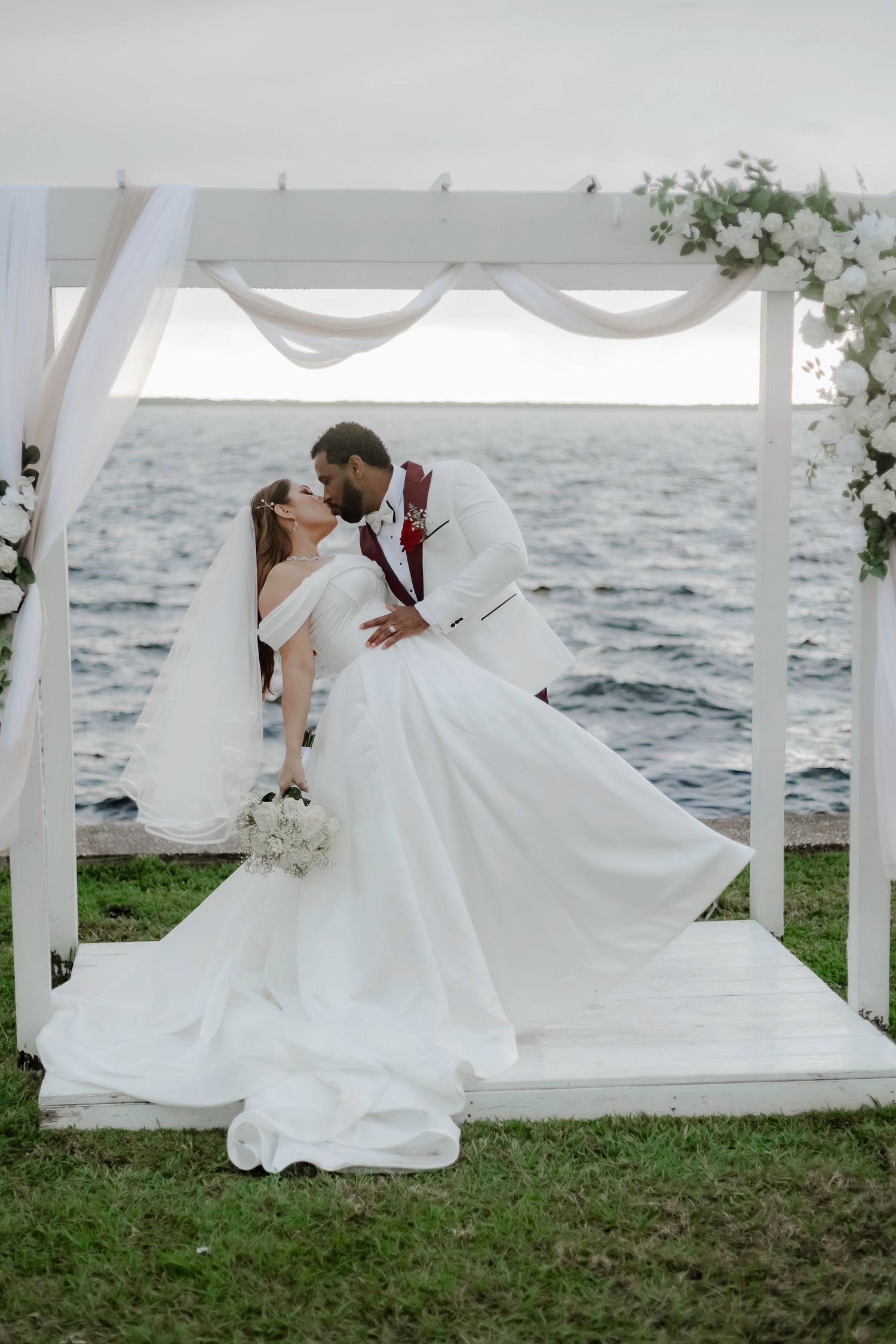 A couple shares an intimate moment under a white wedding arch by the ocean during their seaside ceremony.