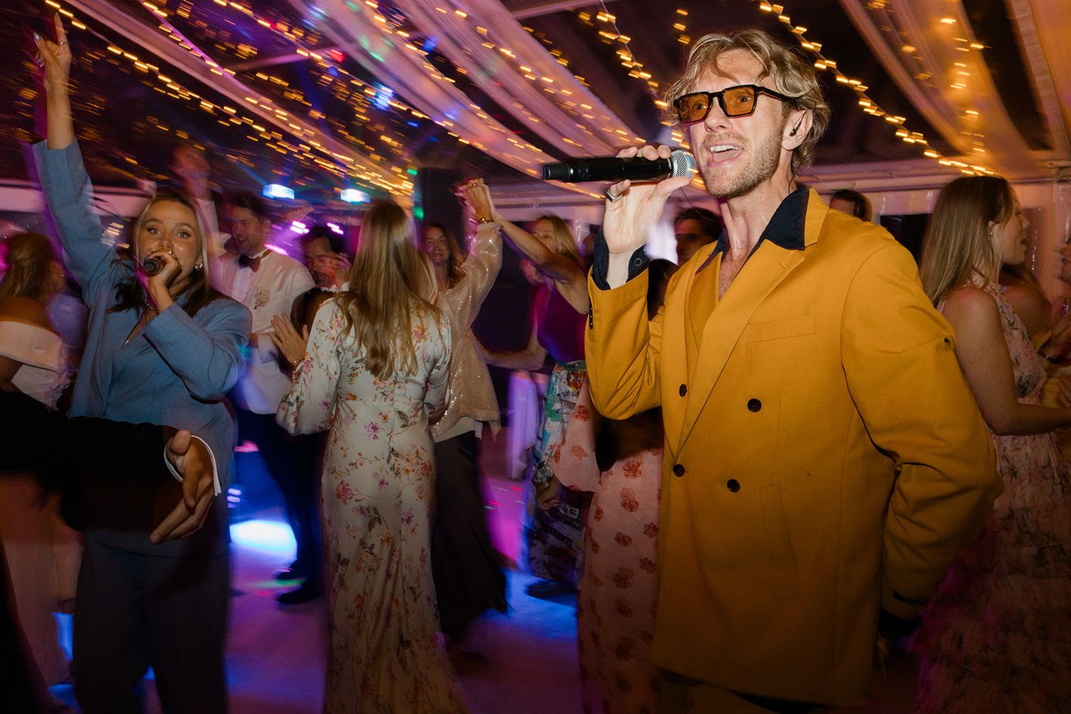 People dancing and celebrating at a lively party with colorful lighting in what appears to be a rustic venue.