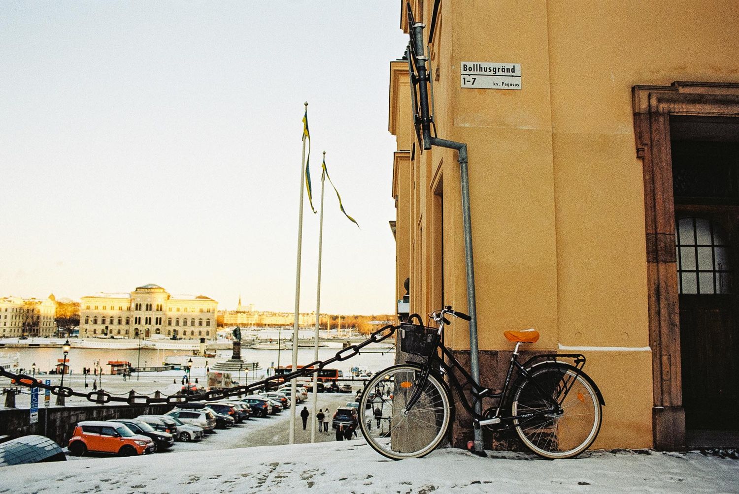 Bicycles parked against yellow walls in a snowy winter urban setting with buildings and parked cars visible.