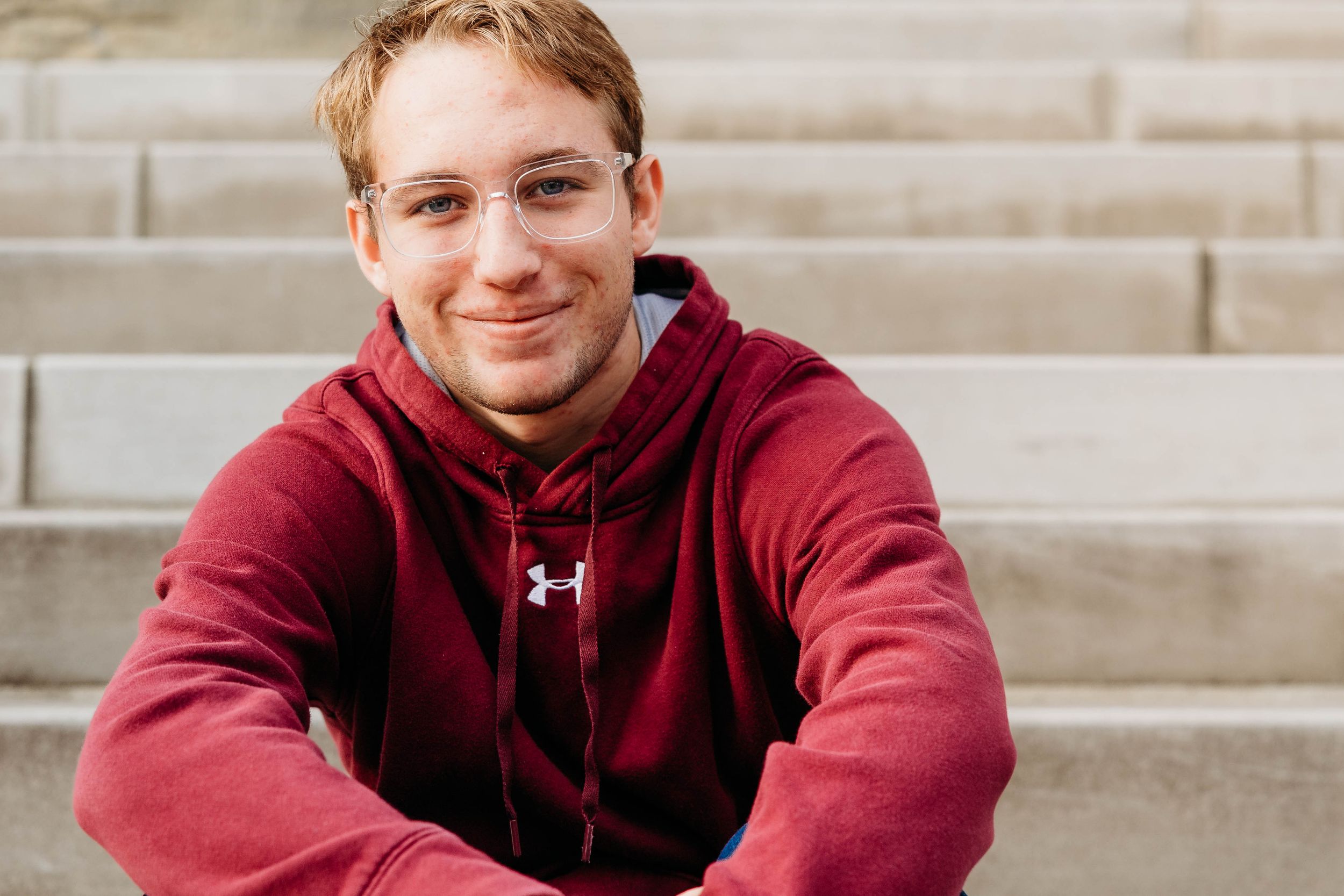 A person wearing a burgundy Under Armour hoodie sits on concrete steps while smiling at the camera.