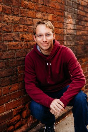 A person in a burgundy hoodie sits against a rustic brick wall, smiling warmly in a casual outdoor portrait series.