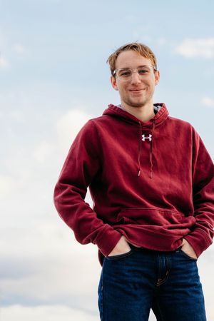 Person in a burgundy hoodie and blue jeans stands outdoors against a cloudy sky background.