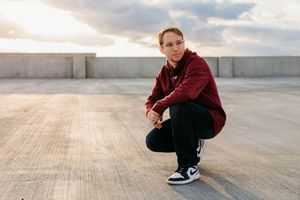 A person in a burgundy sweater and black pants crouches on a concrete rooftop at sunset.