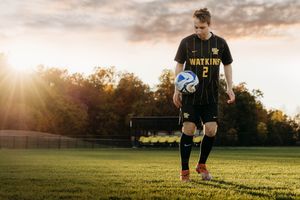 Soccer player dribbling ball on grass field at sunset with golden sunlight creating dramatic silhouette effect.