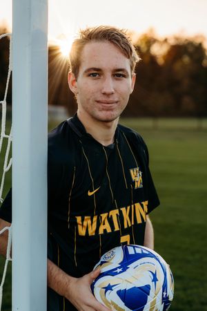Soccer player in black and yellow striped jersey holds a blue ball at sunset on a grassy field.