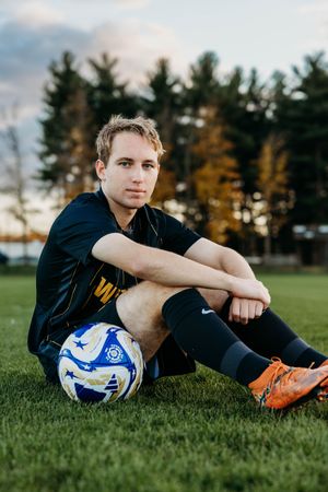 Soccer player rests on grass field with blue and white ball during golden hour with pine trees in background.
