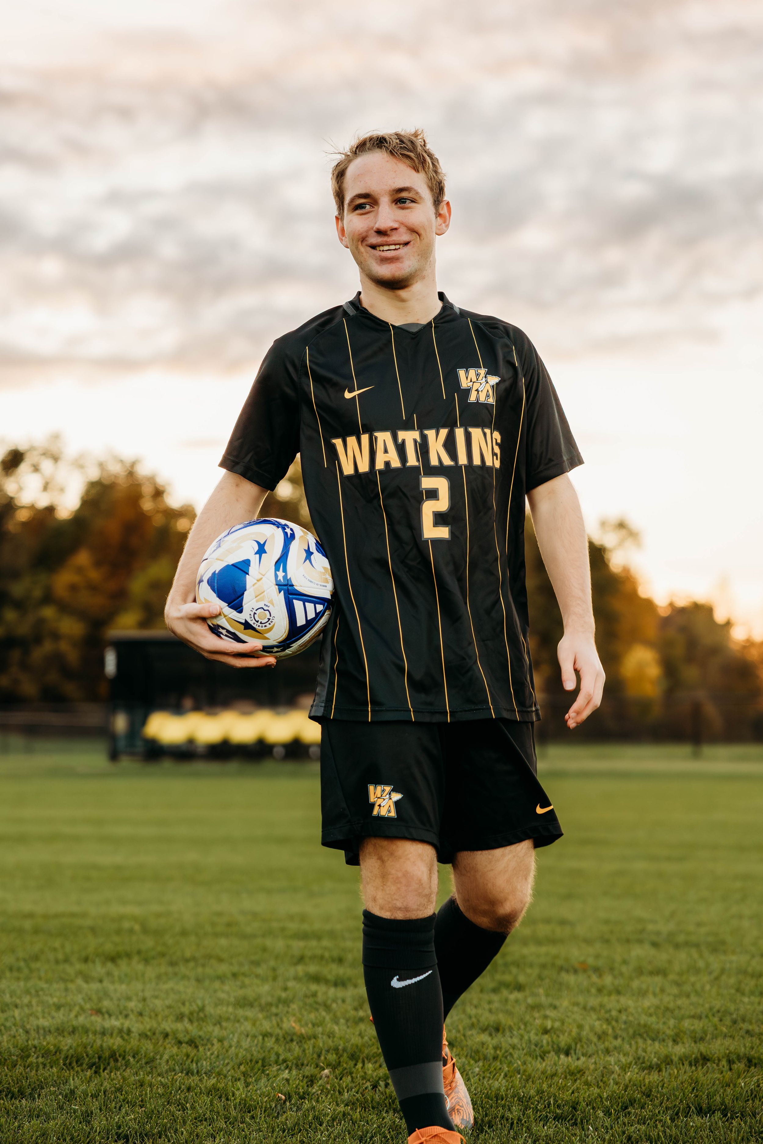 Soccer player in black and gold striped uniform holds blue and white ball on grass field at sunset.