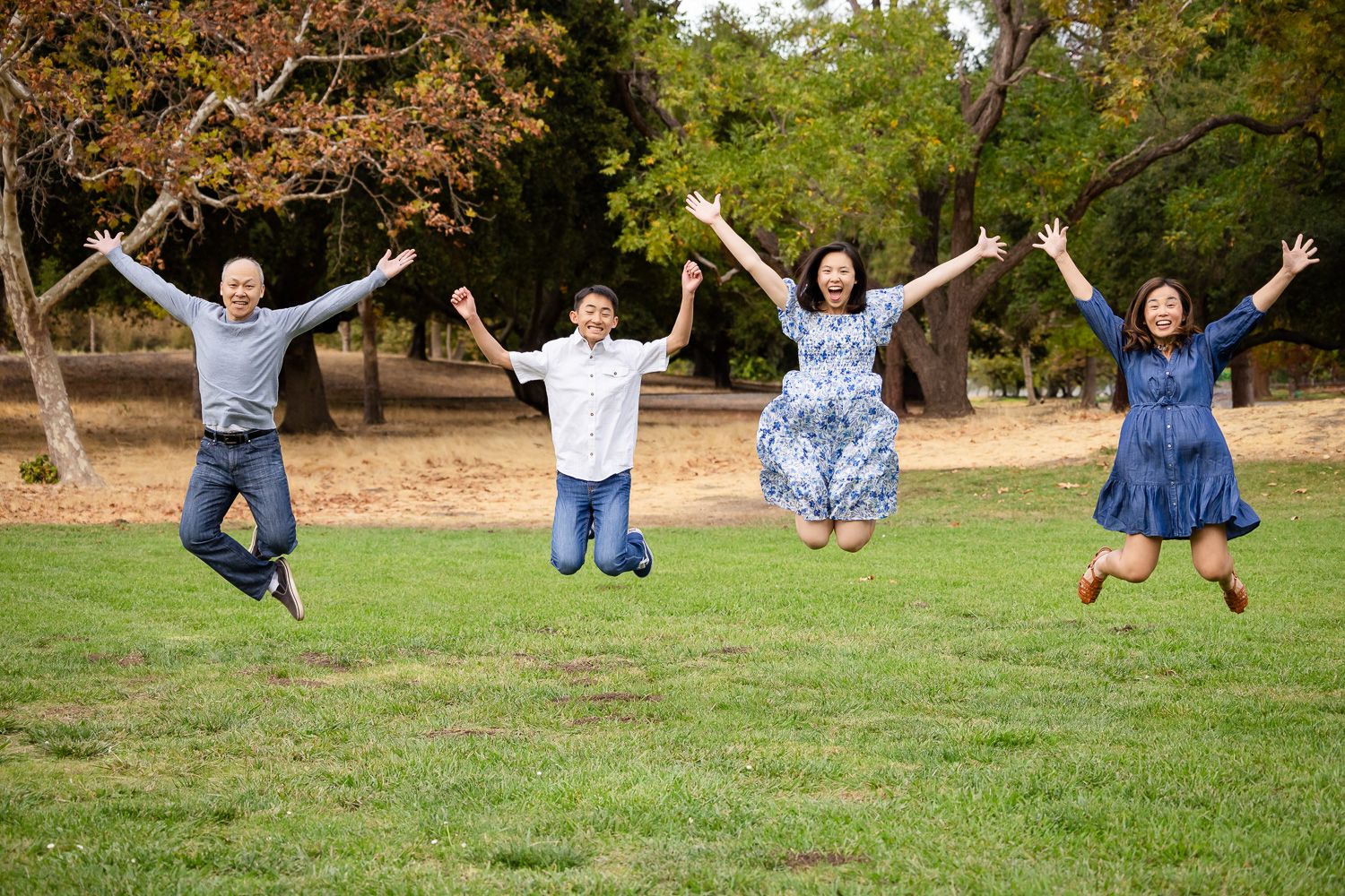 A Palo Alto family jumps joyfully together on grass in a park setting with trees in the background.