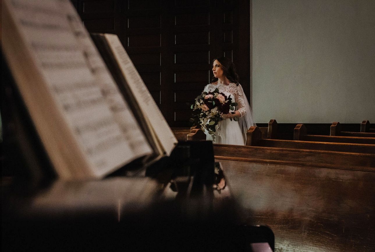 A bride stands in a dimly lit church sanctuary near a piano with sheet music in the foreground.
