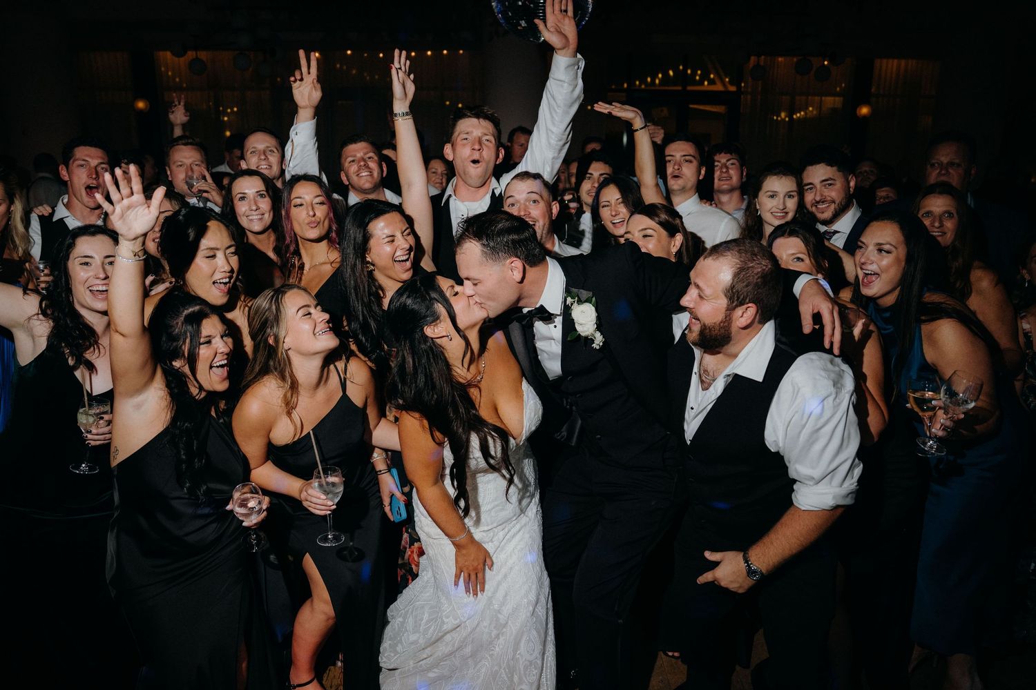 Wedding guests dance and celebrate together on a dark dance floor with their hands raised in excitement.