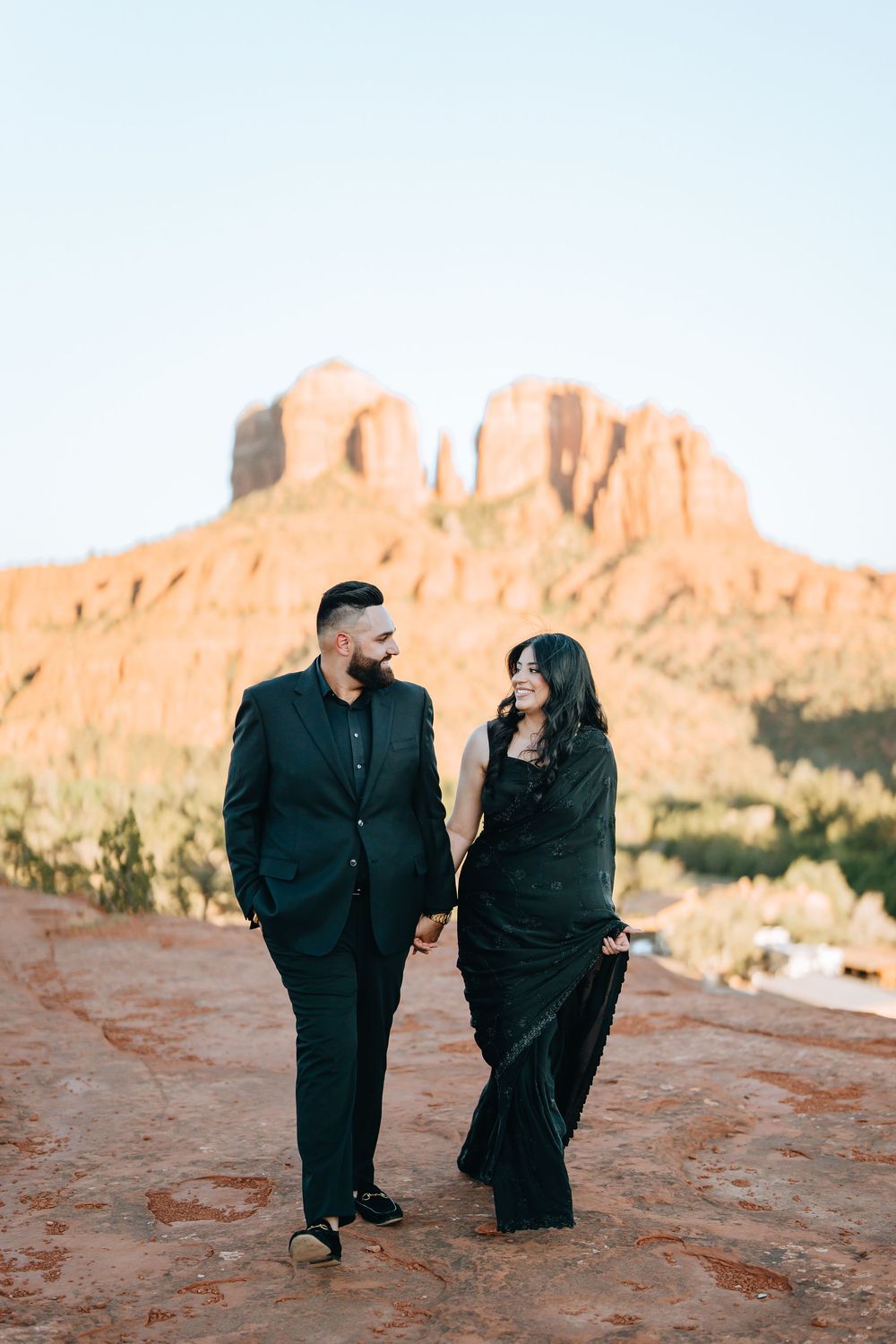Couple walking together on red desert terrain with towering rock formations behind them.