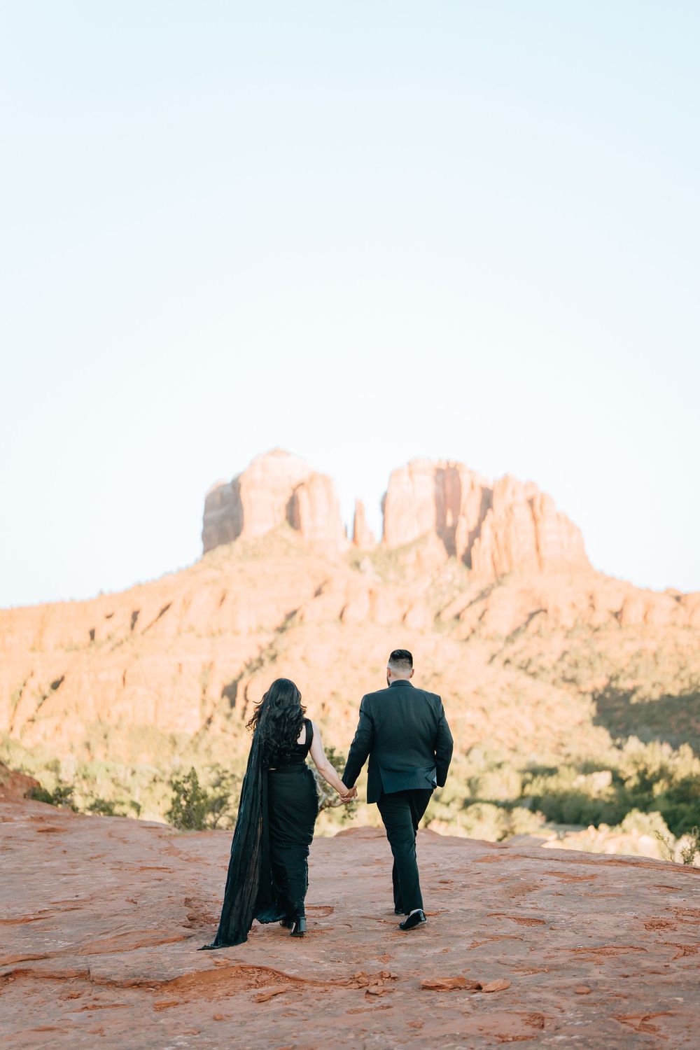 A couple dressed in black shares a romantic moment against stunning red rock formations in the desert landscape.