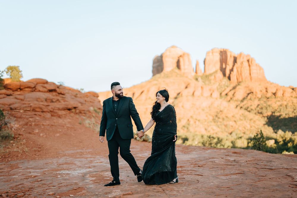 Two people in dark attire walking together on red desert rocks with towering cliffs in the background.