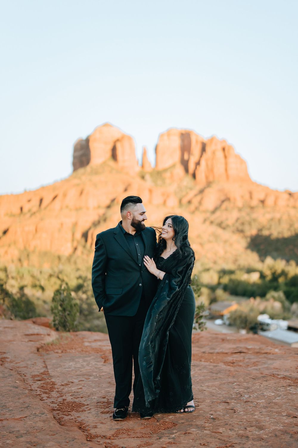 Couple in black clothing sharing tender moments on red rock terrain with desert cliffs at sunset.