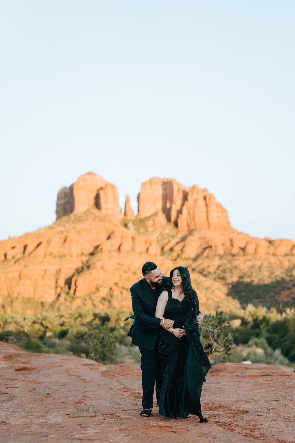 Two figures in dark clothing embrace on rustic red desert rocks with mountains in the distance.