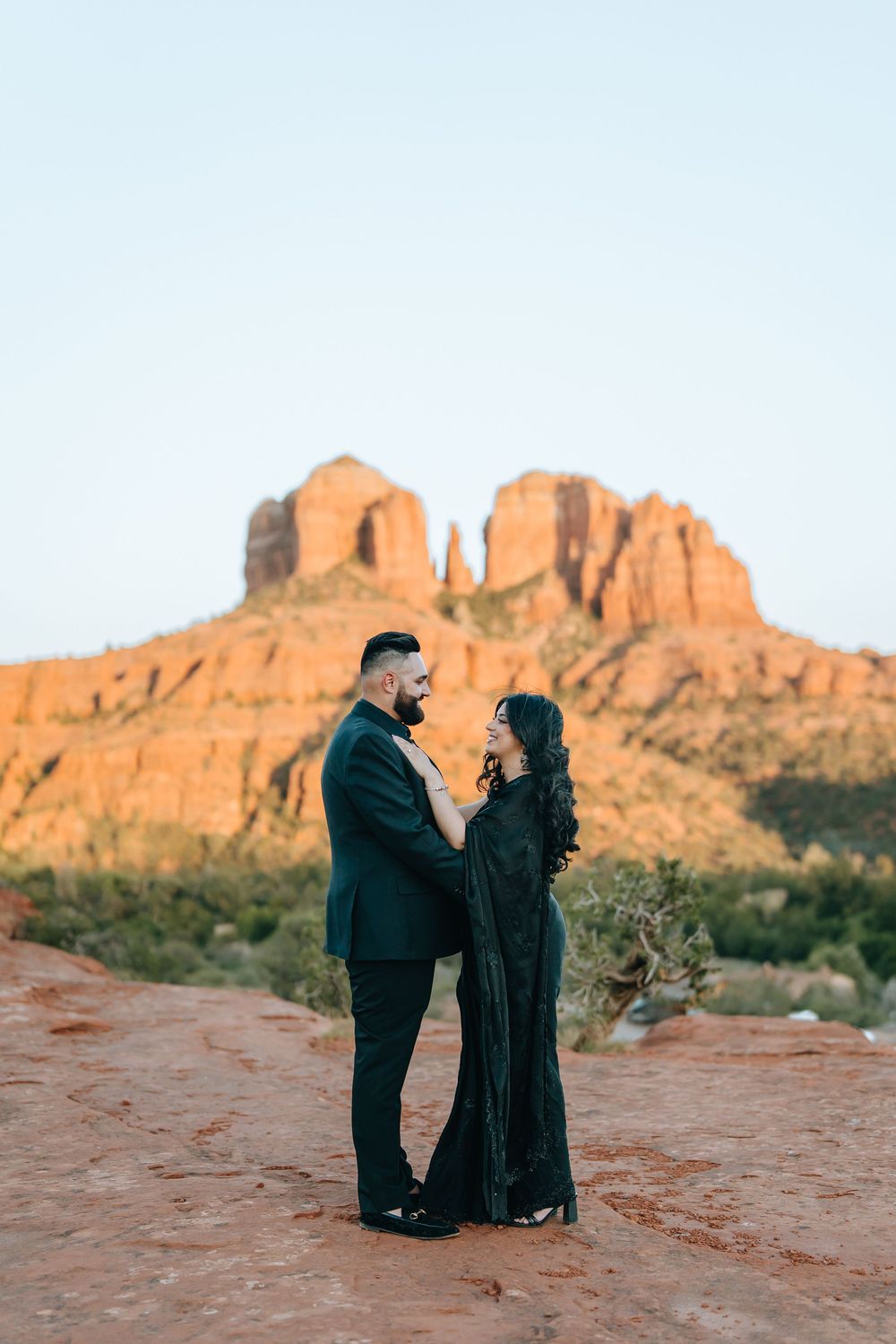 Two people in dark clothing pose romantically on red rocks with desert mountains behind them.