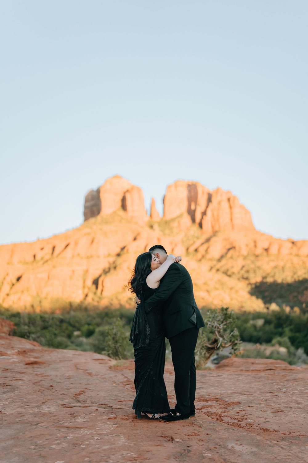 Couple in black attire standing against majestic red rock formations at sunset.